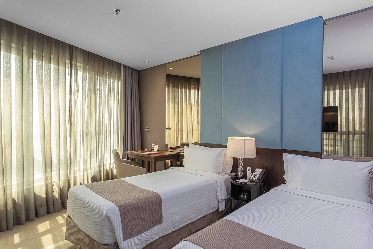 Quarto de hotel com duas camas, uma escrivaninha, uma cadeira, um abajur e um telefone.
