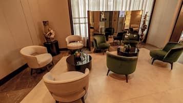 Eine Lobby mit einer Sitzecke, ausgestattet mit mehreren Sesseln und einem Couchtisch, einer Statue auf einem Sockel und einem großen Spiegel, der den Raum reflektiert.