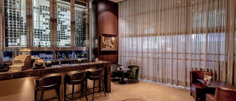 A bar area with a wine rack, bar stools, and a counter. There are two round tables with chairs, and a large window with curtains.