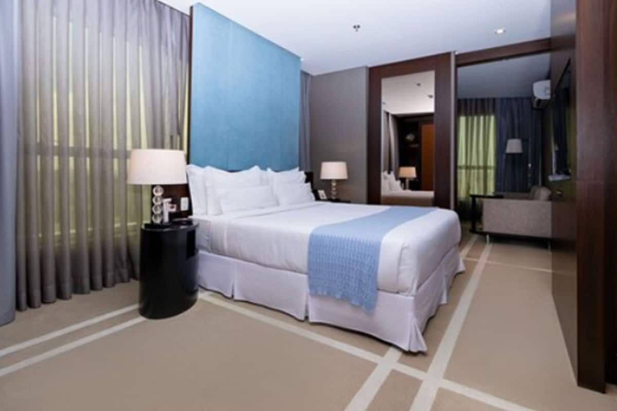 Quarto de hotel com uma cama grande, duas mesas de cabeceira com abajures, uma parede azul de destaque e vista para outro quarto através de uma porta aberta.