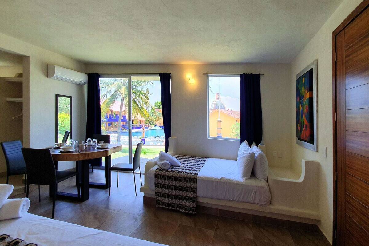 Quarto de hotel com cama, escrivaninha, cadeira e vista para uma piscina e palmeiras.