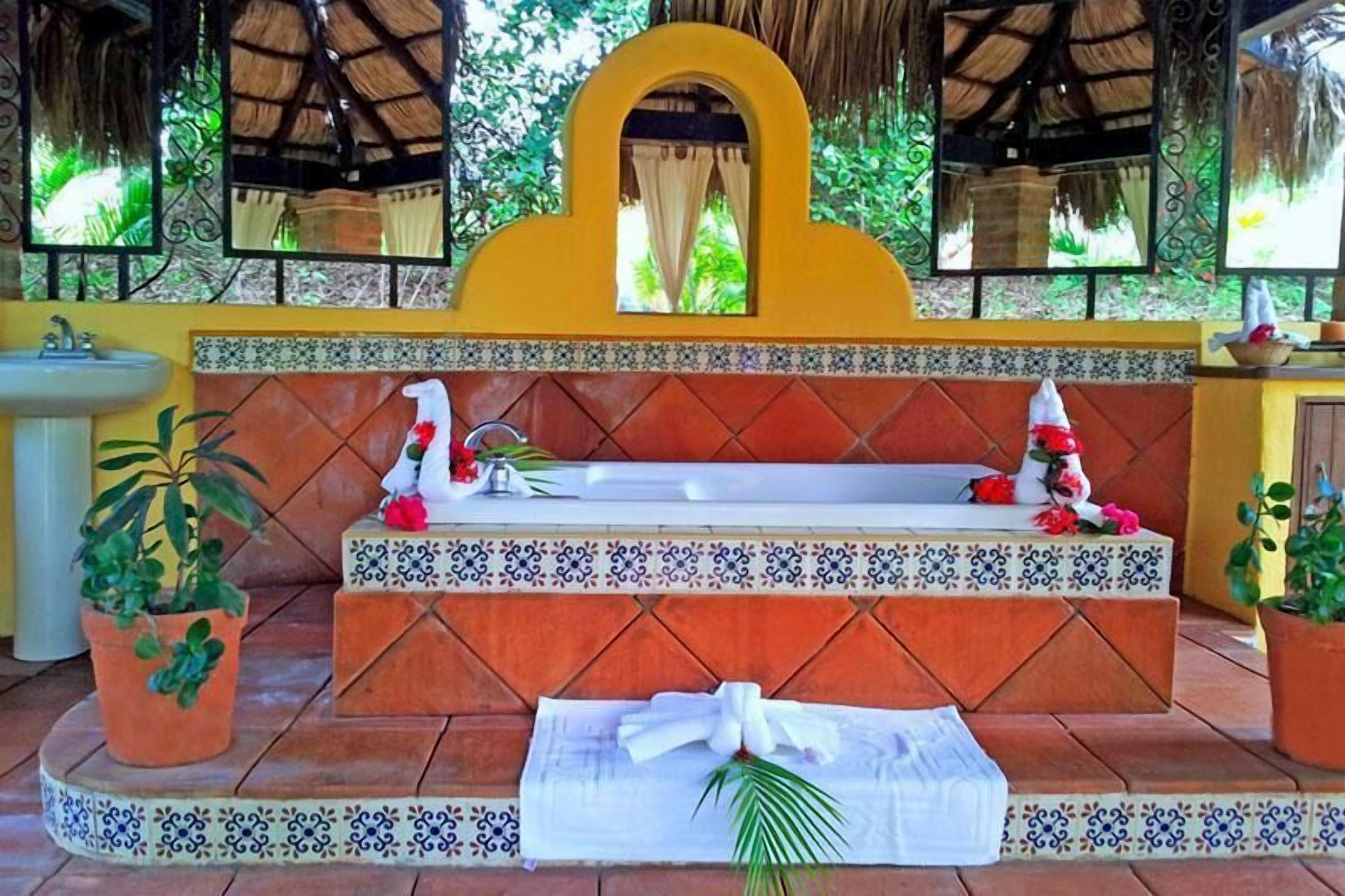 A bathroom with a bathtub, a sink, and a mirror. There are potted plants and a towel on the floor.