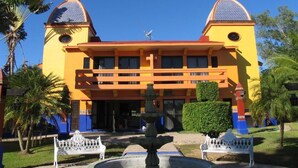 A yellow building with two towers, a fountain, and a well-maintained garden with benches.