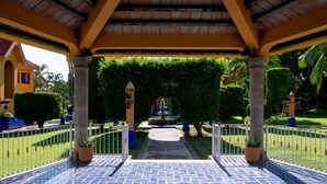 A covered walkway with a tiled floor leading to a fountain, surrounded by greenery and a white fence.