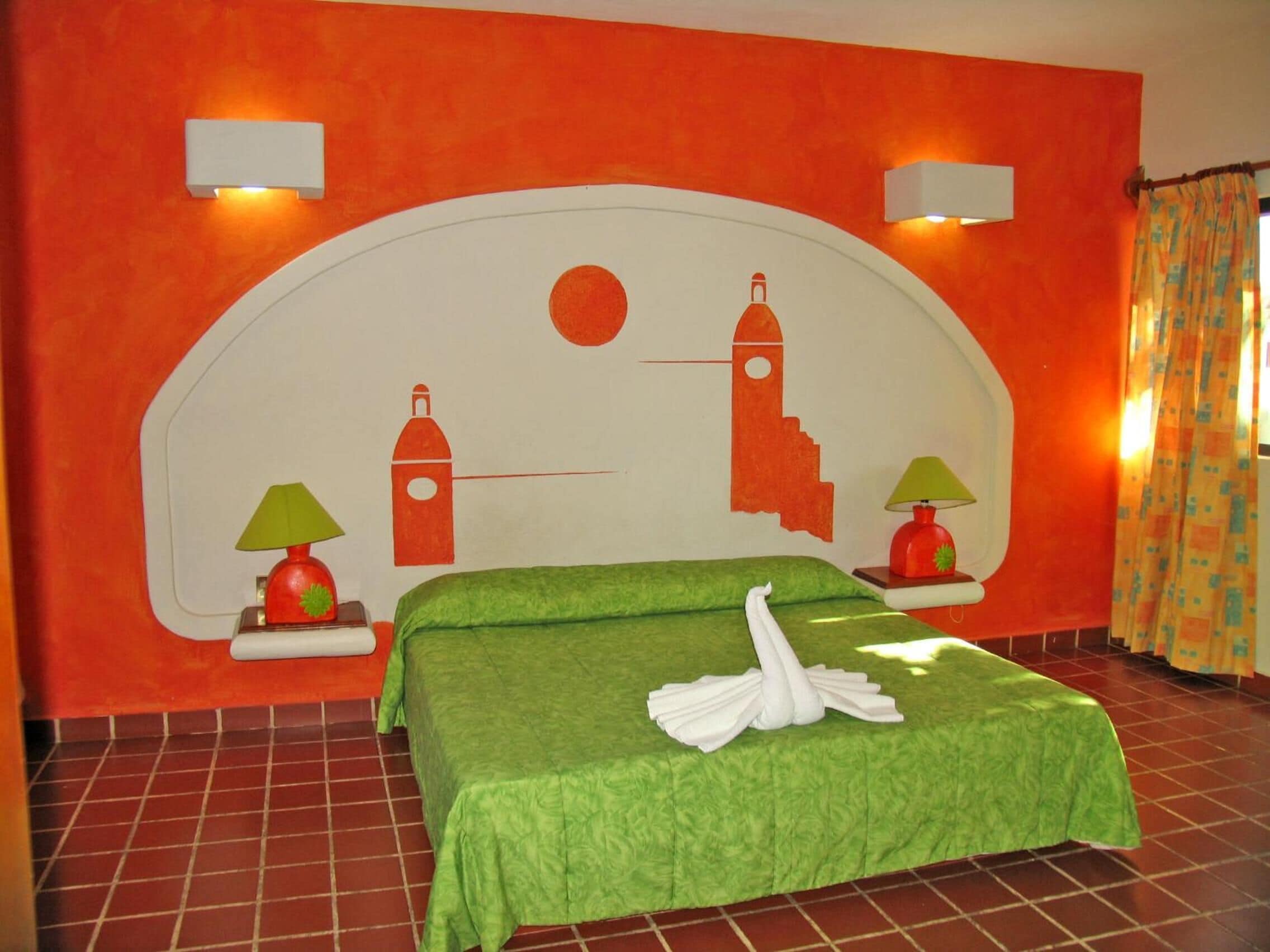 A hotel room with a large bed, green bedding, two wall-mounted lamps, and a mural with a sun and buildings.