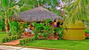 A thatched-roof hut with a small table and chairs, surrounded by lush greenery and a well-maintained garden.