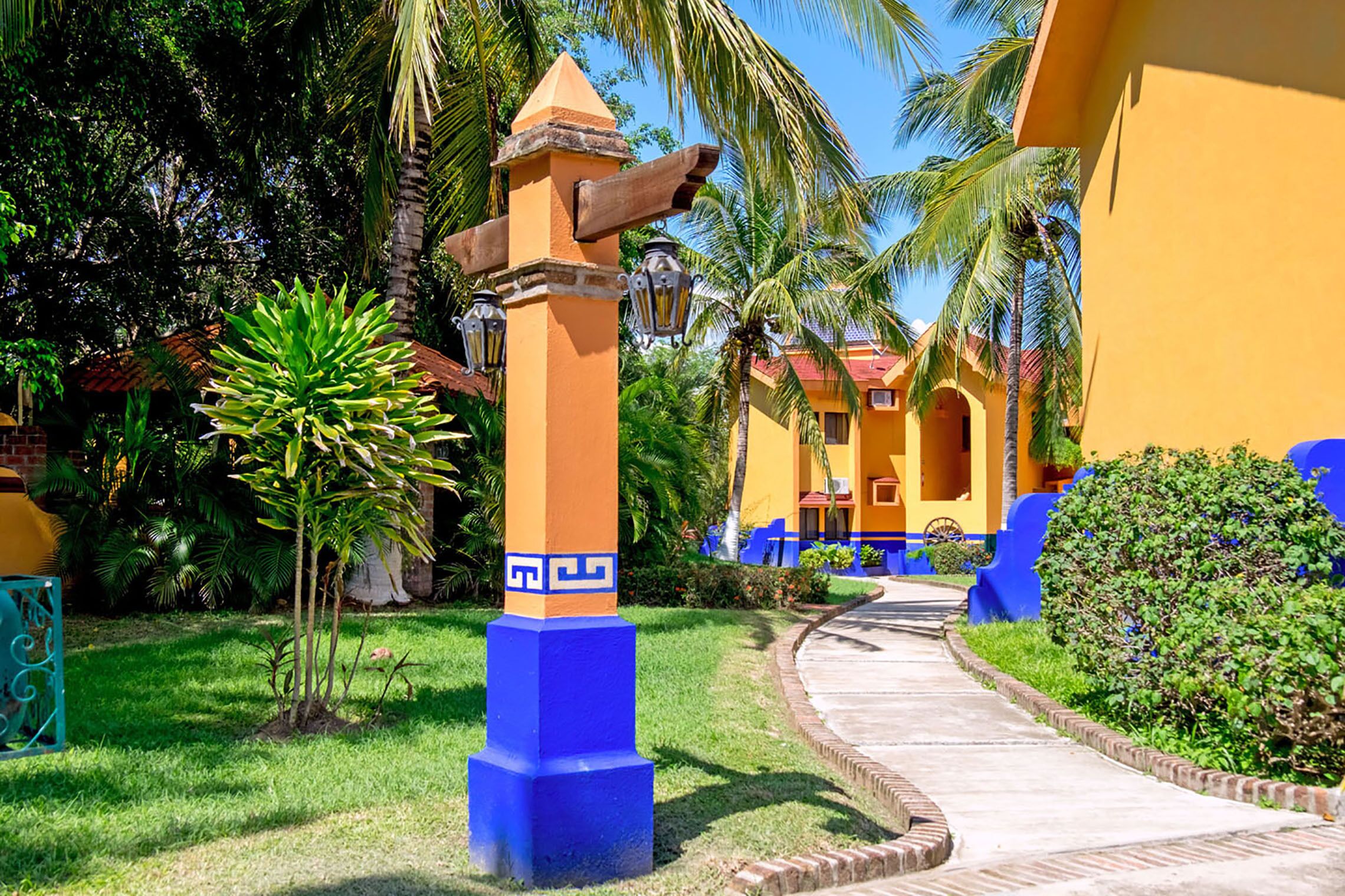 A pathway leading to a building with a yellow facade and blue accents, surrounded by lush greenery and palm trees.
