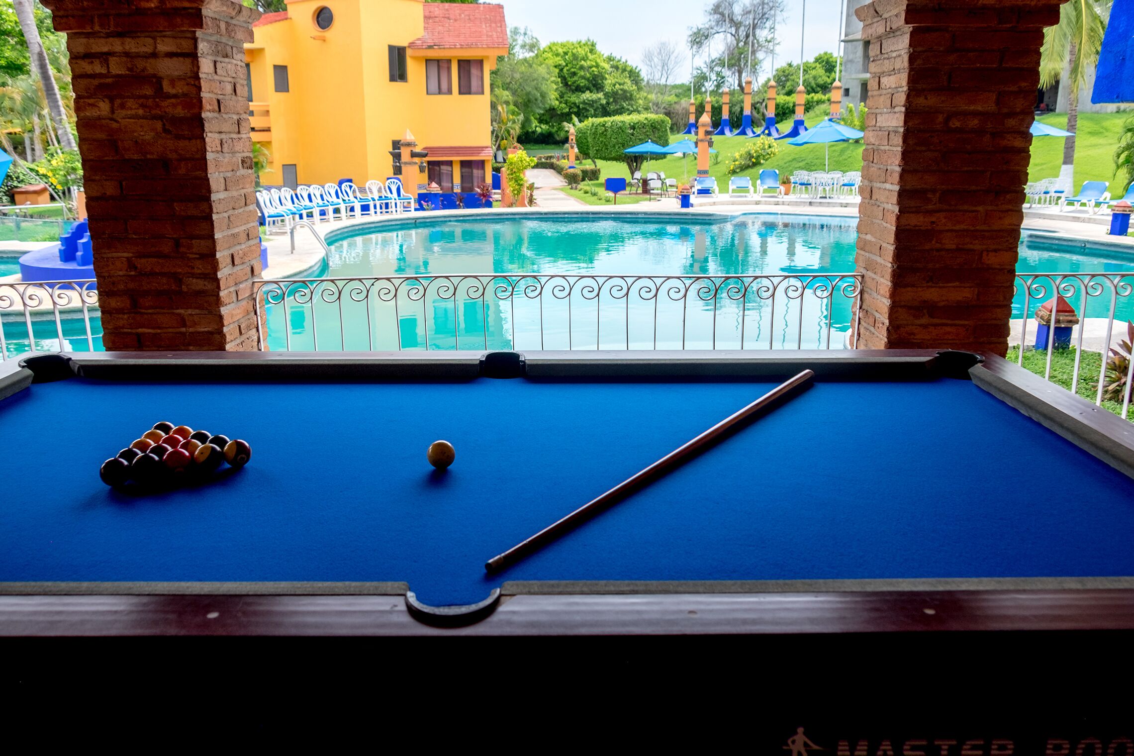 A pool table with balls and a pool cue, overlooking a swimming pool with lounge chairs and umbrellas.