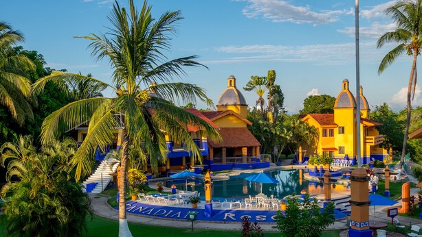 A resort with a swimming pool, palm trees, and a building with a yellow facade.