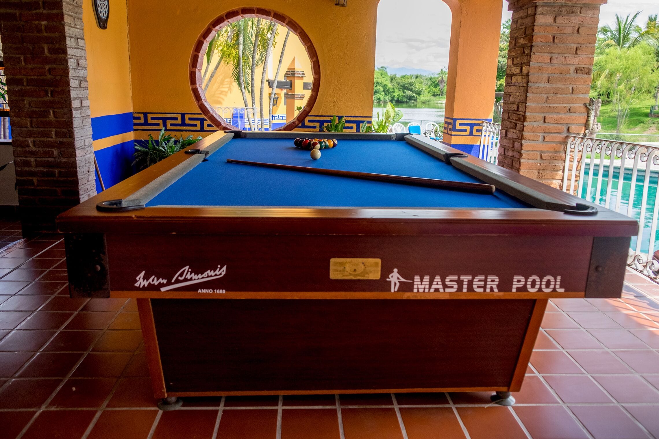 Una mesa de billar con el rótulo "MASTER POOL", con vista a una piscina y palmeras al fondo.