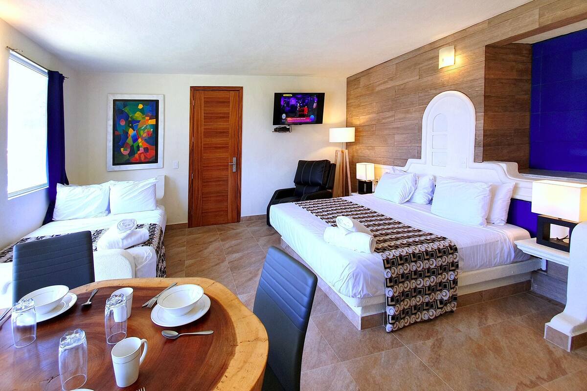 Quarto de hotel com cama, escrivaninha com cadeira, mesa de jantar com cadeiras e televisão fixada na parede.