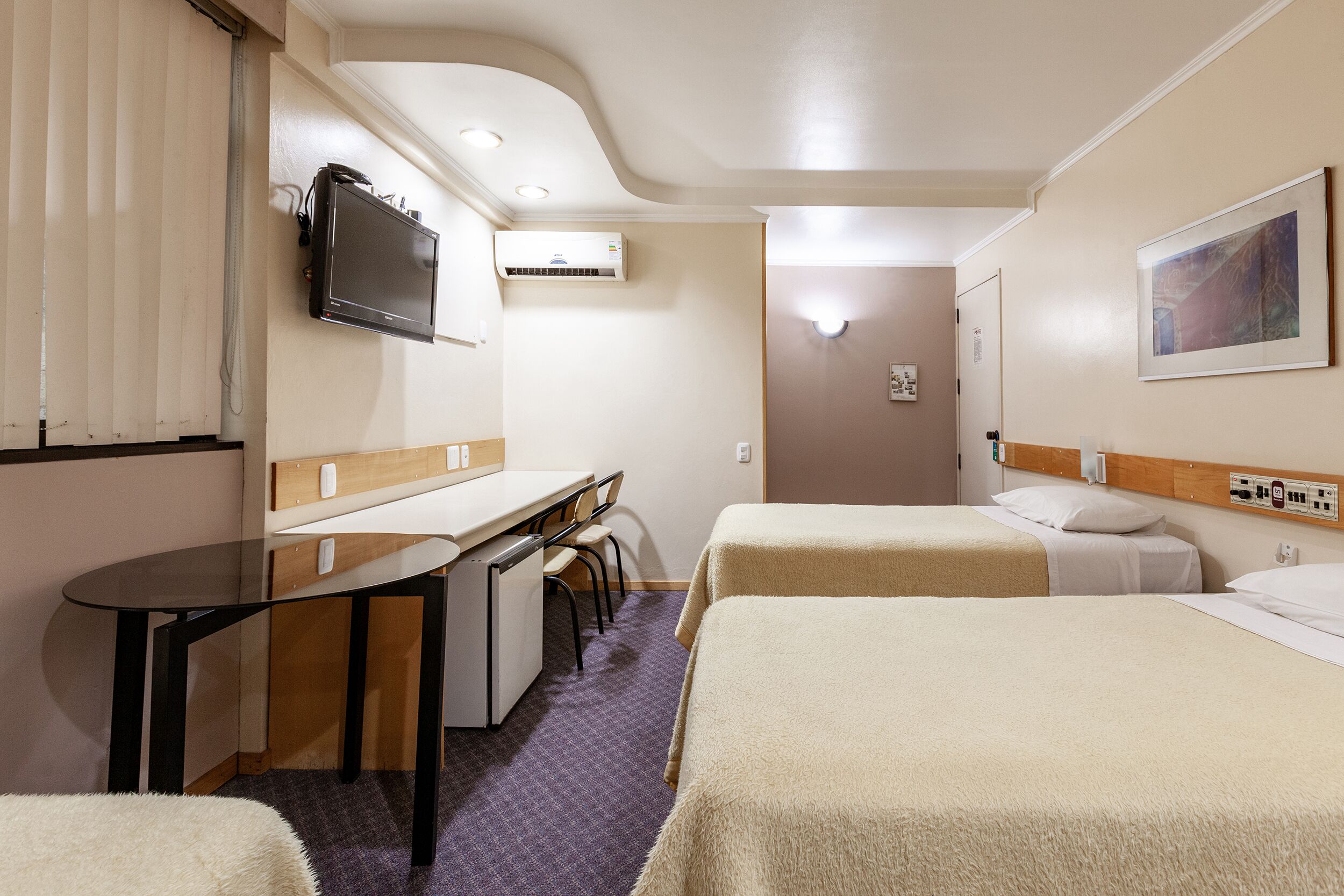 A hotel room with a desk, two beds, a TV, and an air conditioning unit.