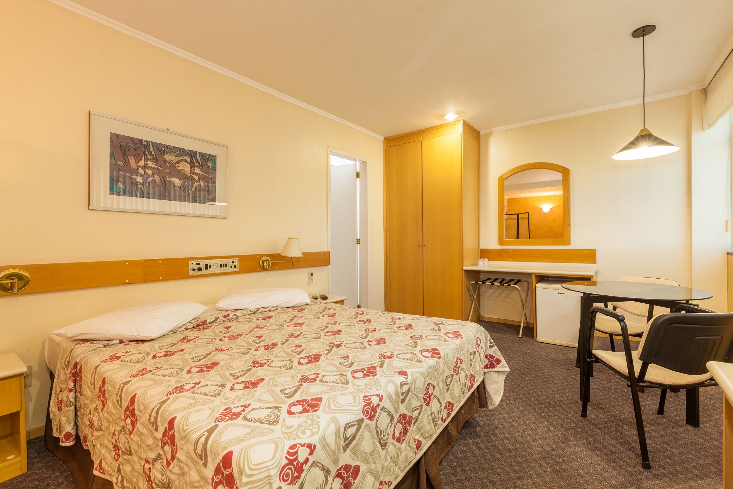 A hotel room with a bed, a desk, a chair, a mirror, and a painting on the wall.