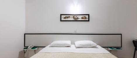 Un dormitorio moderno con una cama grande, ropa de cama blanca, una obra de arte enmarcada sobre la cama y una mesita de noche con un teléfono y un control remoto.
