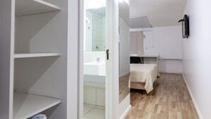 Una habitación pequeña y diáfana con kitchenette, una cama con colcha blanca y un baño visible a través de una puerta abierta.