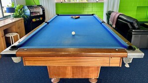Een pooltafel met een blauwe viltkleed, omringd door groene bakstenen muren, een raam met witte gordijnen en een poolkeu-rek.