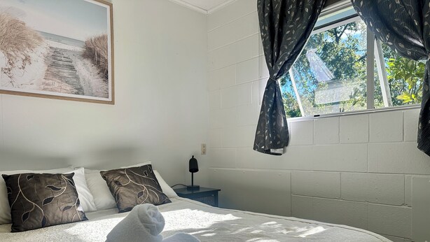 Een net opgemaakt bed met een wit-bruin dekbed, twee kussens en een opgerolde witte handdoek. Op de muur hangt een schilderij van een strandgezicht, en er is een raam met gordijnen te zien.