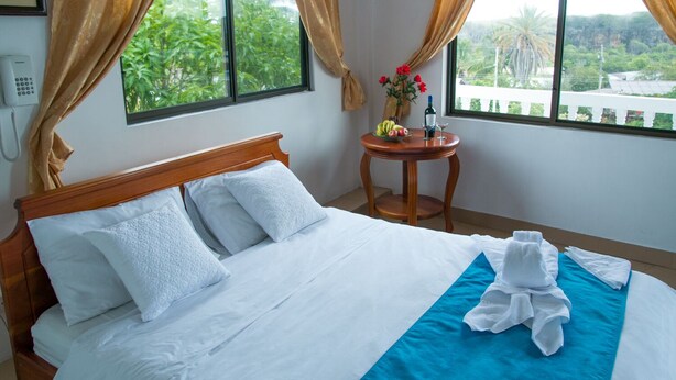 Una cama bien hecha con sábanas blancas y un detalle en azul, una pequeña mesa redonda con un florero y una botella, y vistas a la vegetación a través de amplias ventanas.