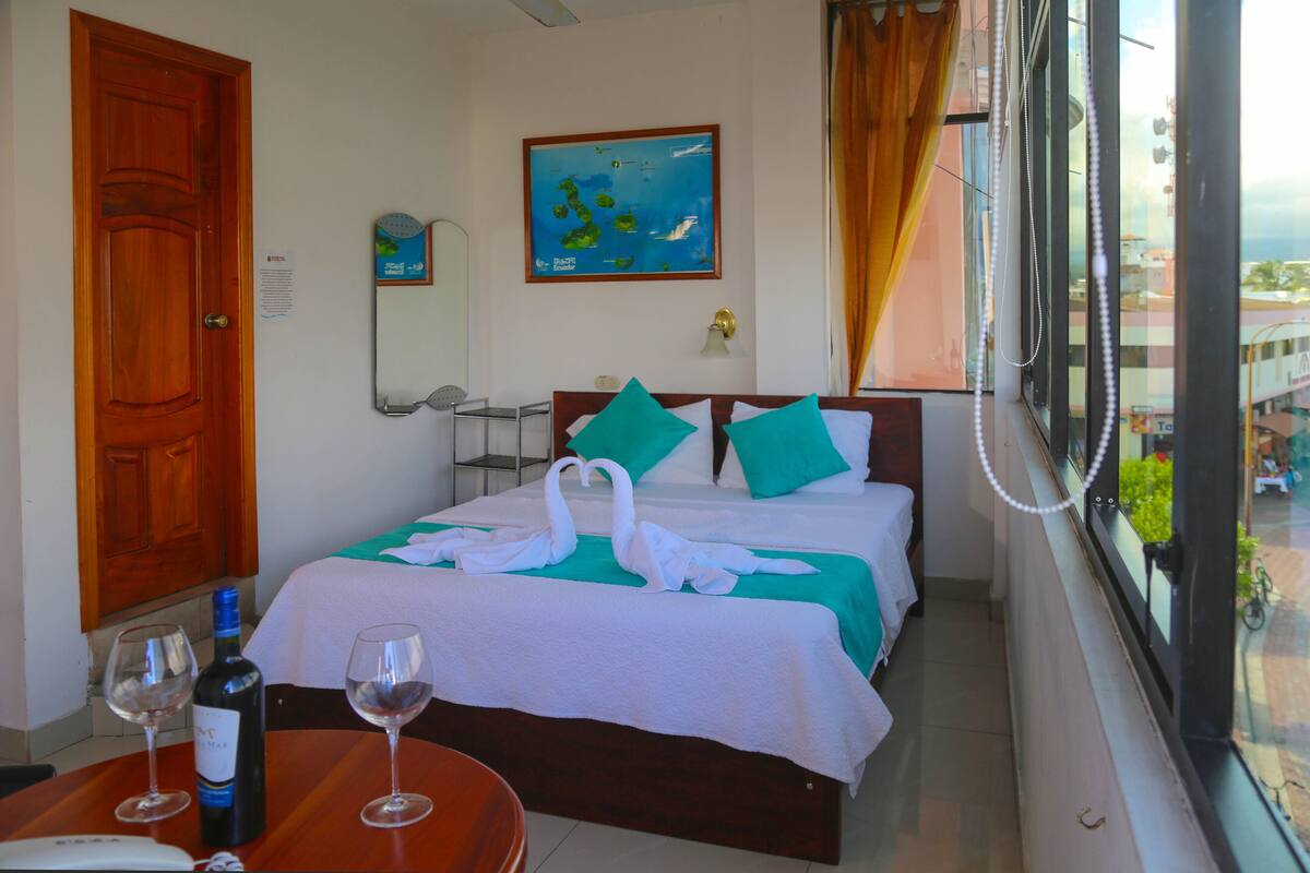 Quarto de hotel com duas camas, uma porta de madeira, um mapa na parede, uma janela com vista e uma mesa com uma garrafa e copos.