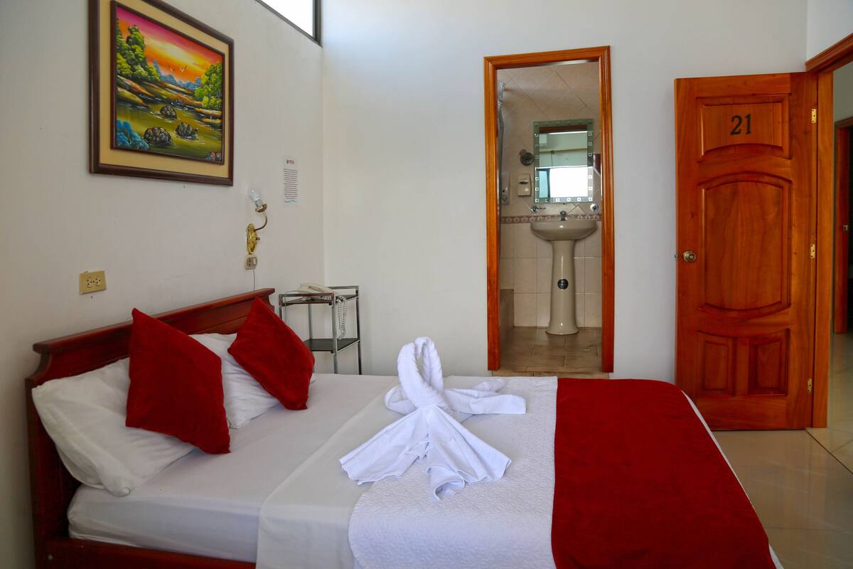 Quarto de hotel com uma cama, almofadas vermelhas, uma toalha branca, um quadro e um banheiro visível por uma porta aberta.