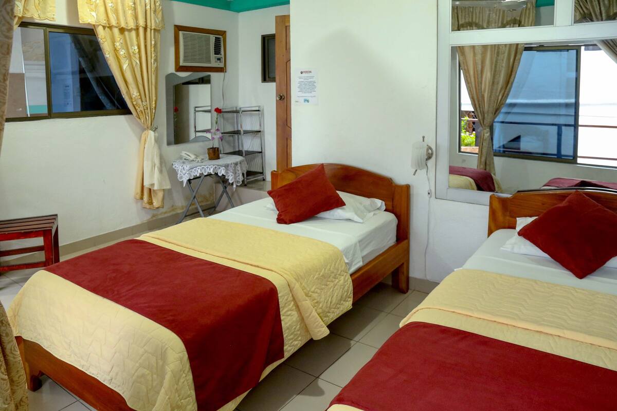 Quarto de hotel com duas camas, cada uma com colcha vermelha e bege, cabeceira de madeira e janela com cortinas.