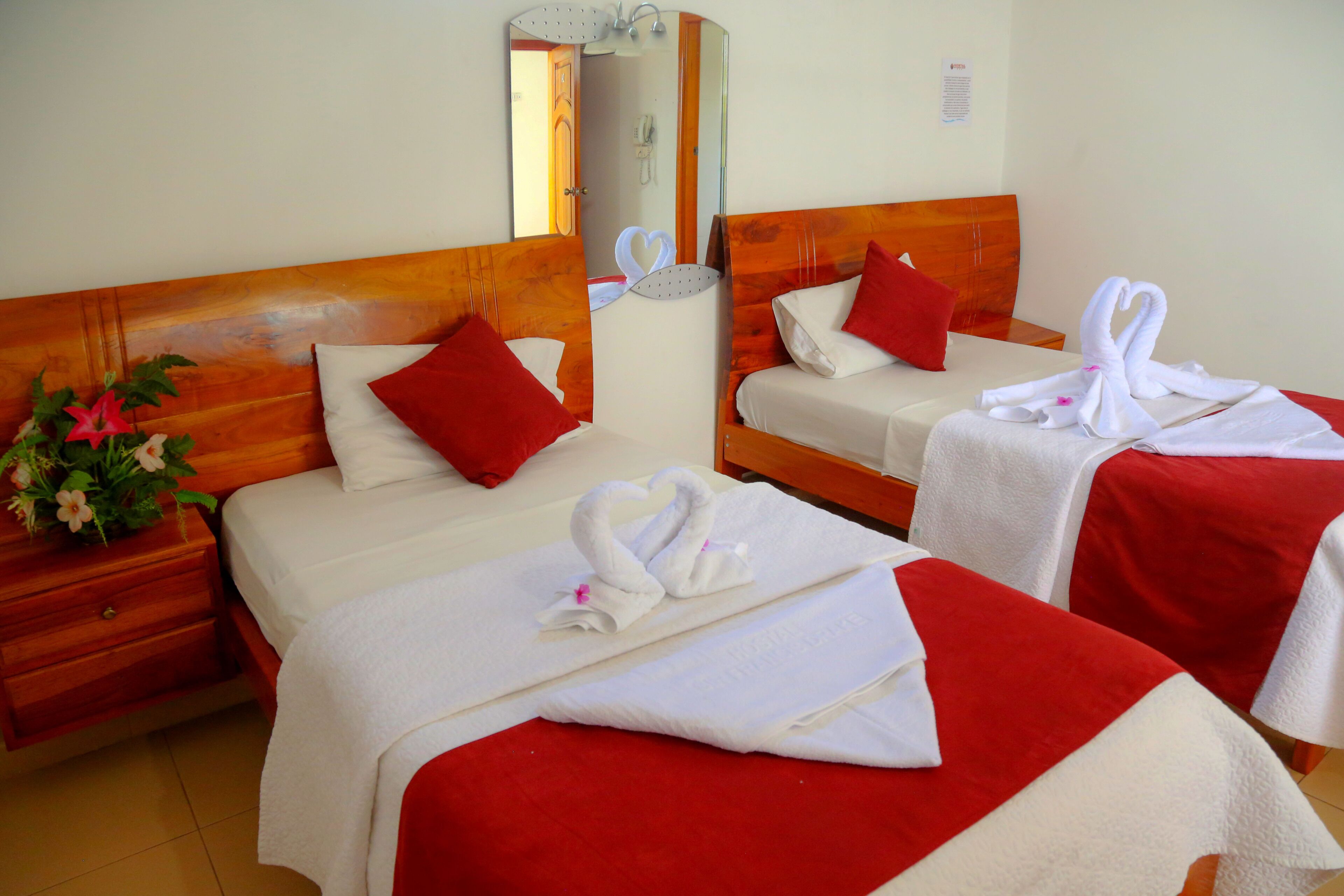 Une chambre d’hôtel avec deux lits, chacun agrémenté d’un oreiller rouge et d’une serviette blanche en forme de cygne. On y trouve une tête de lit en bois, une table de chevet avec une plante et un miroir au-dessus des lits.
