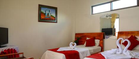 A hotel room with two beds, each with red bedding and white towels arranged in swan shapes. There is a painting on the wall, a ceiling fan, and a window with curtains.