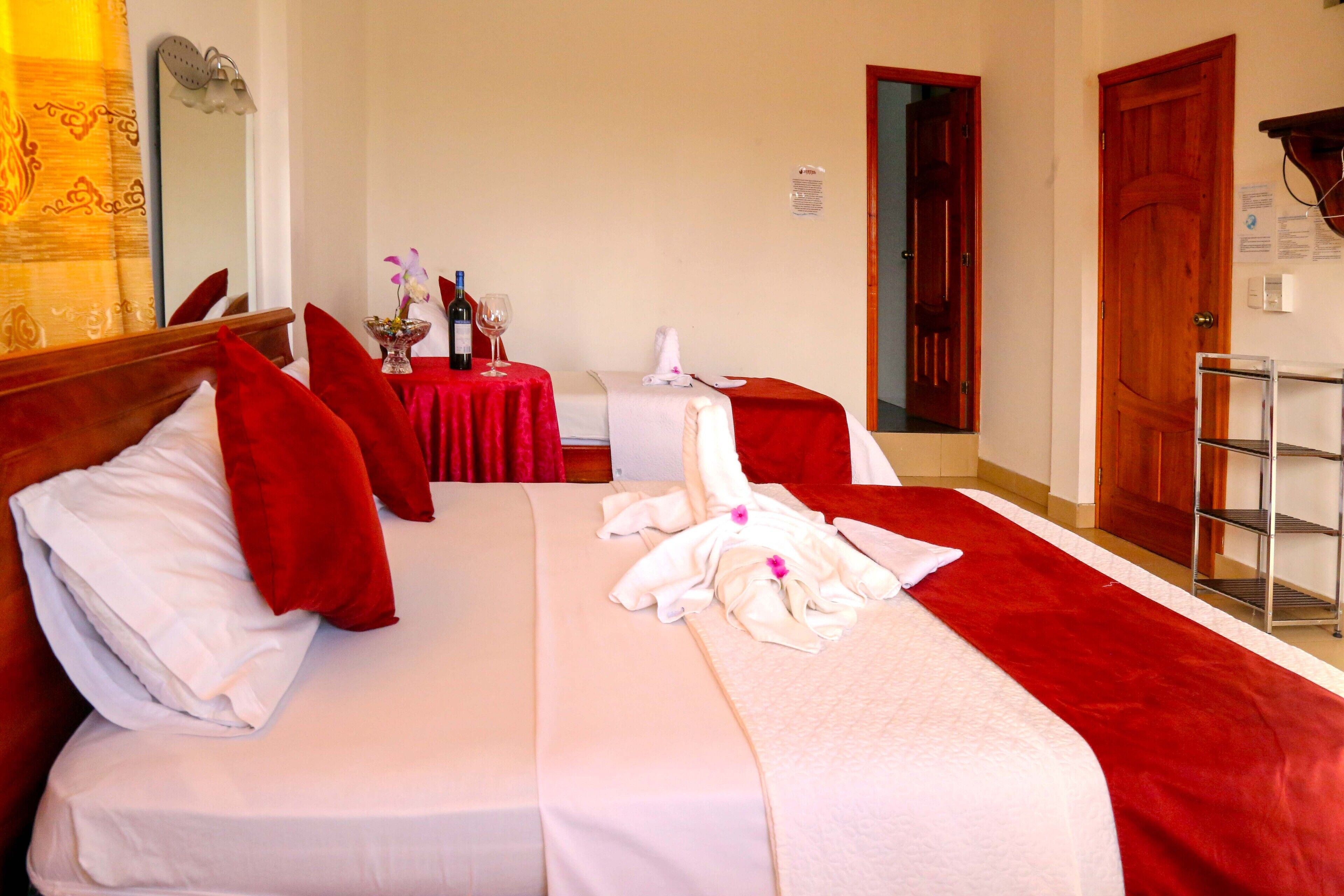 A hotel room with two beds, red pillows, a red bedspread, a small table with a vase and wine glasses, and a wooden door.