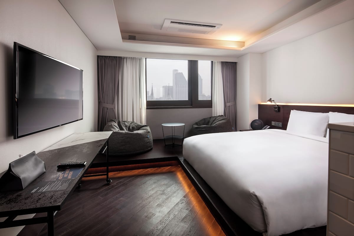 A modern hotel room with a large bed, a flat-screen TV mounted on the wall, a small round table, and a seating area with a sofa and armchair.
