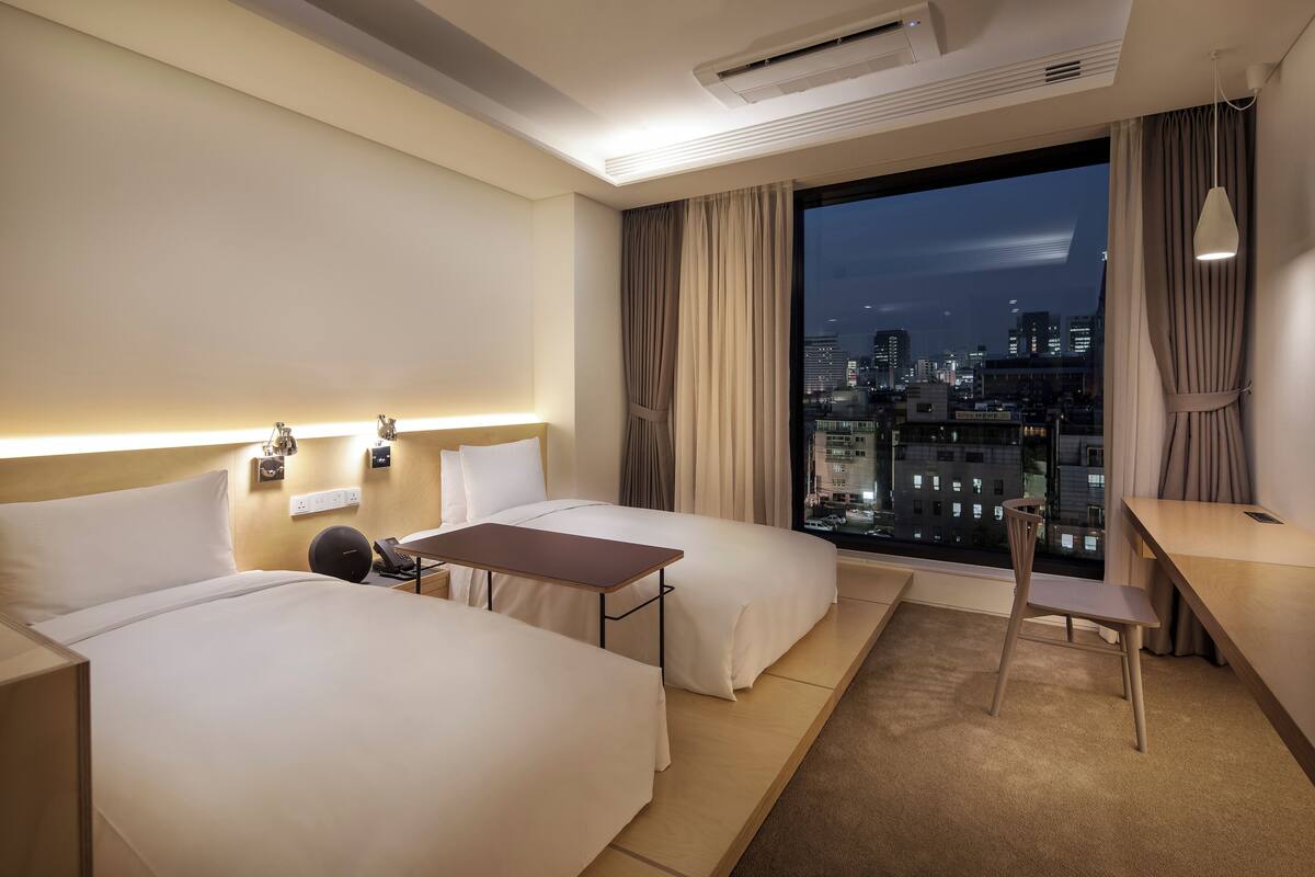A hotel room with two beds, a desk, a chair, a phone, and a city view.