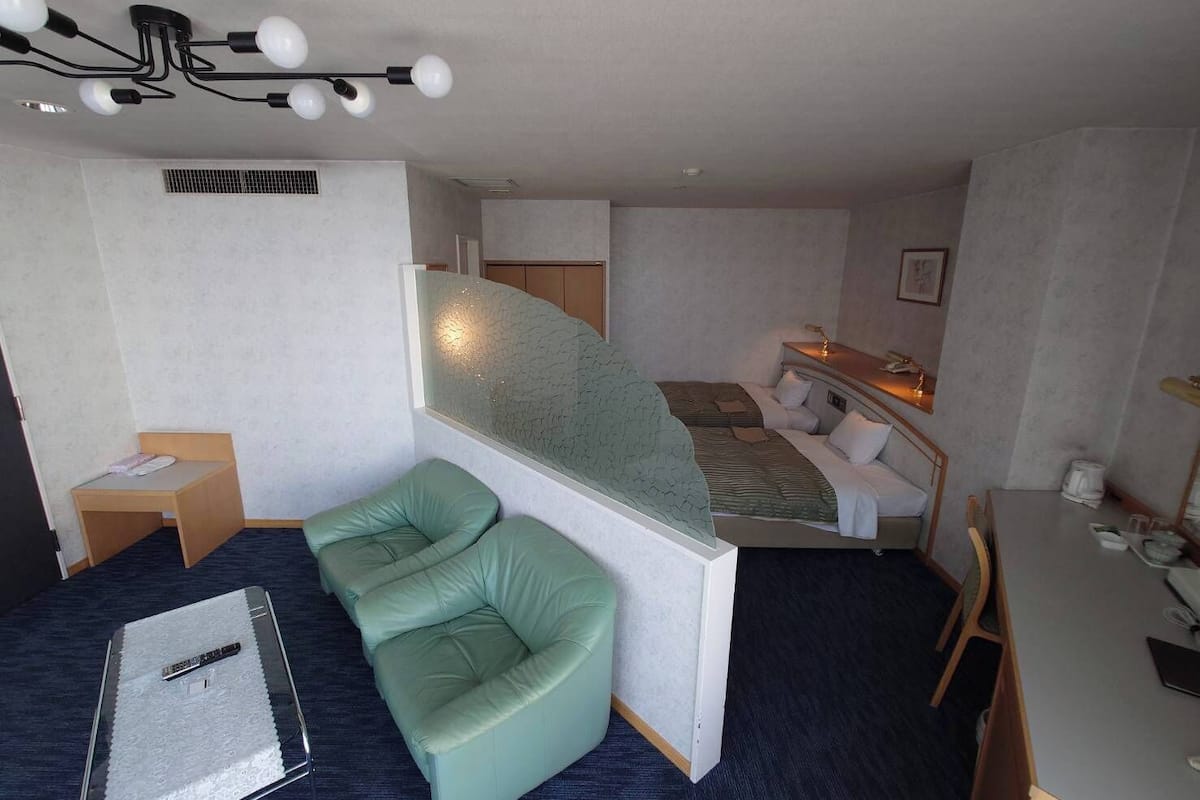 A compact hotel room with a bed, a green cushioned seating area, a desk, and a small table.