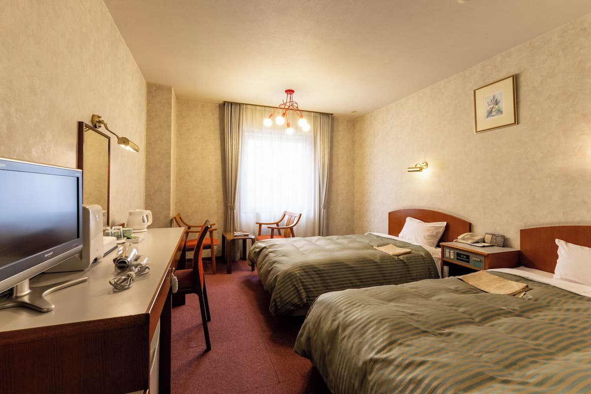 A hotel room with two beds, a desk with a computer, a chair, a television, and a window with curtains.
