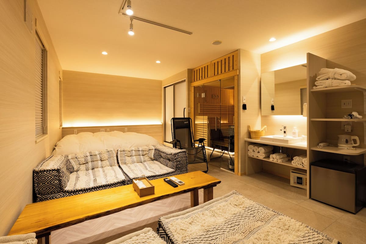A hotel room with a bed, a bedside table with a remote control, a wooden bench, a large mirror, a built-in shower, and a towel rack.