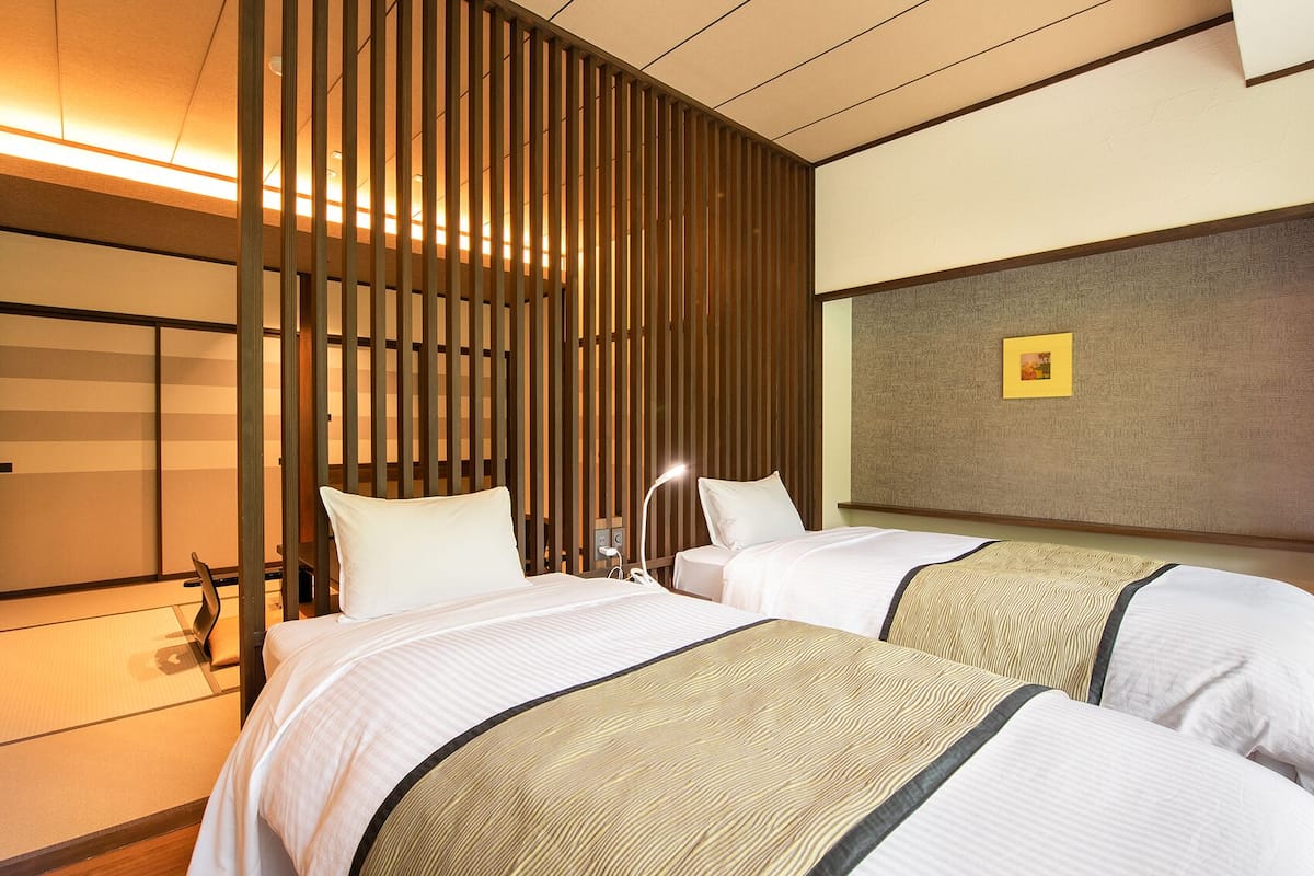 A hotel room with two beds, a wooden slat partition, a bedside table with a lamp, and a framed picture on the wall.