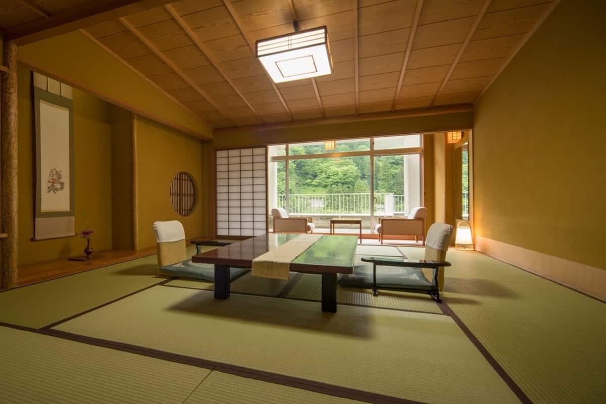 A tatami mat room with a low table, chairs, a shoji screen, and a view of greenery outside.