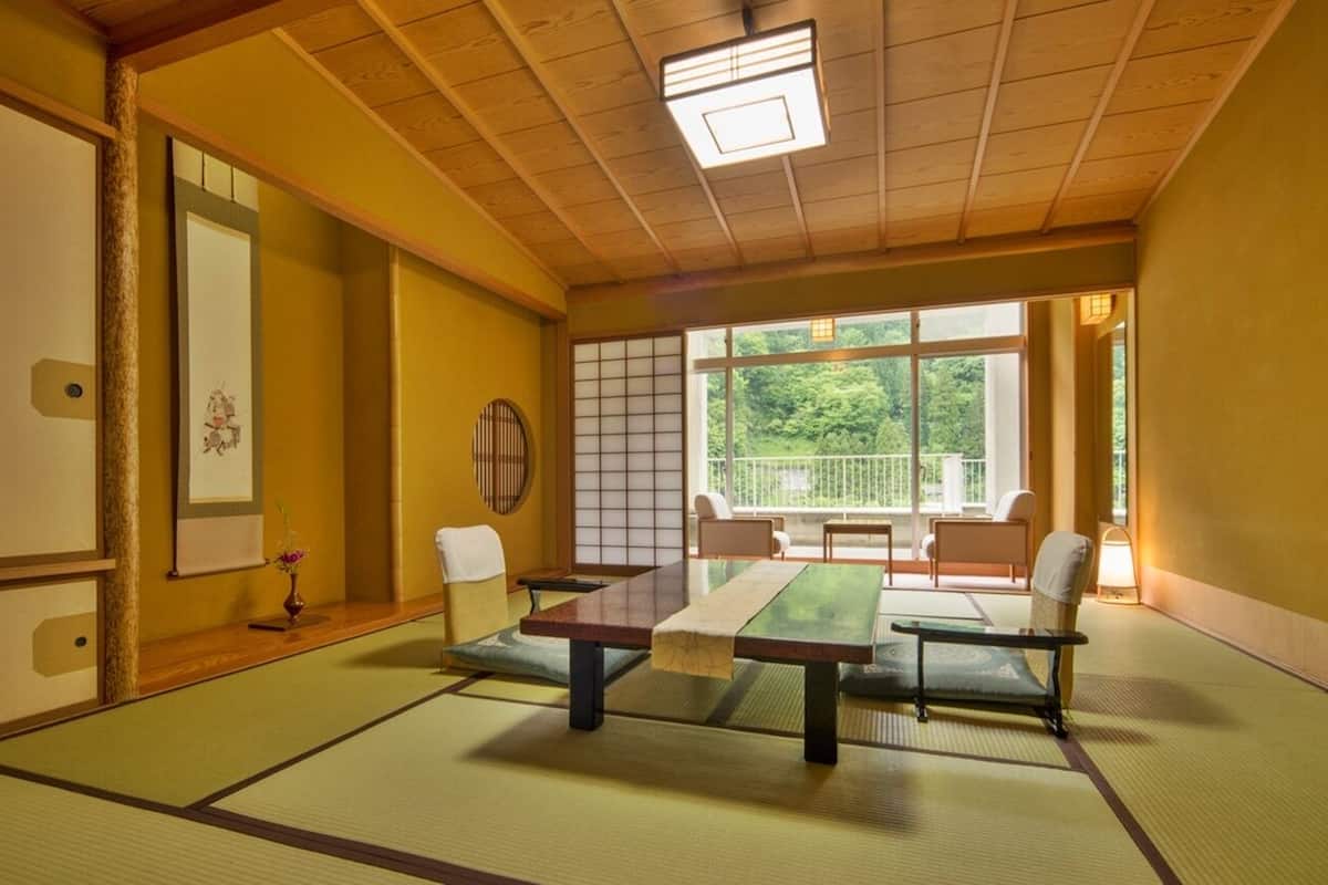 A tatami mat room with a low table and chairs, a shoji screen, and a view of greenery outside.