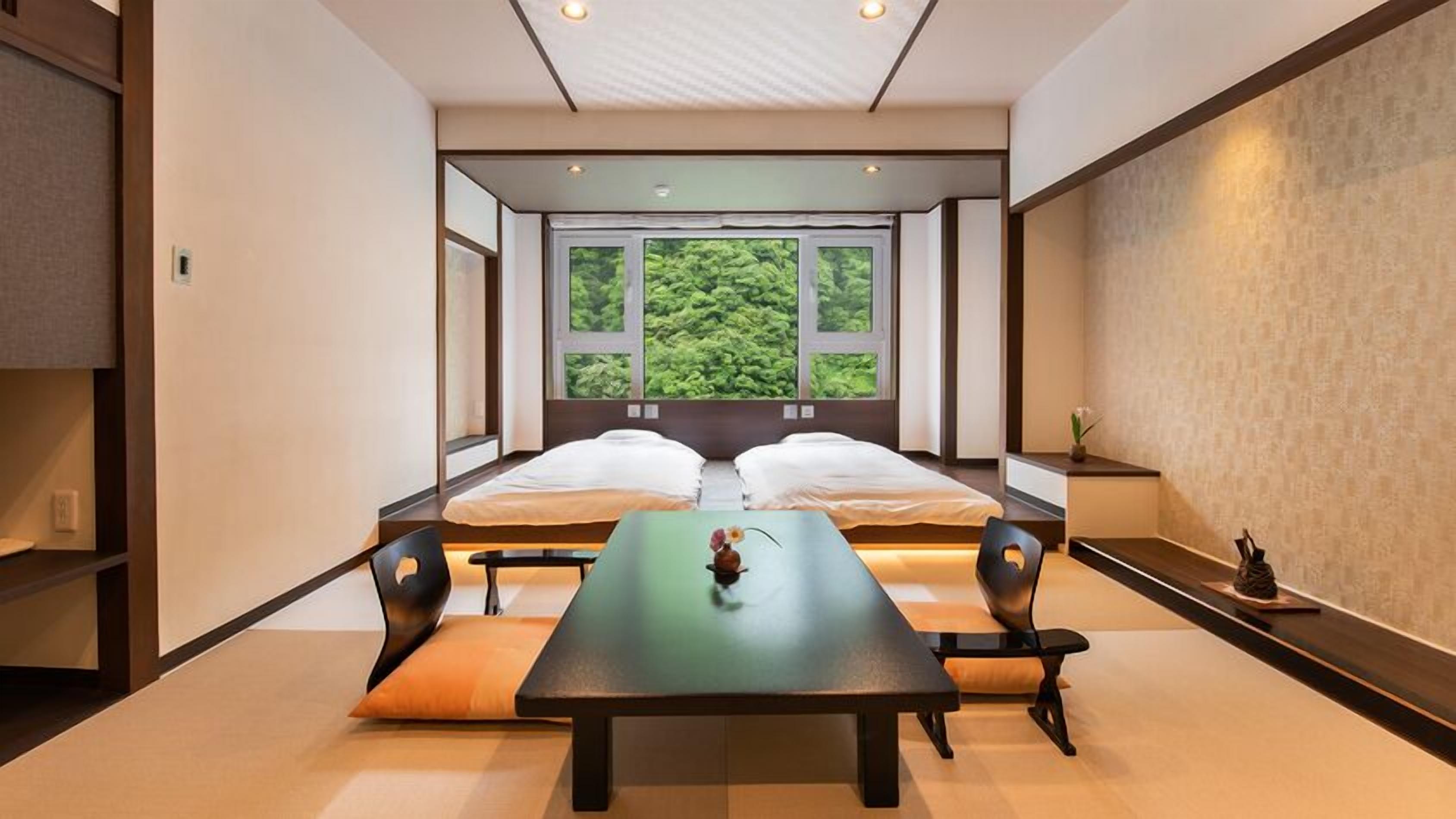 A room with two beds, a table, chairs, and a view of greenery through large windows.