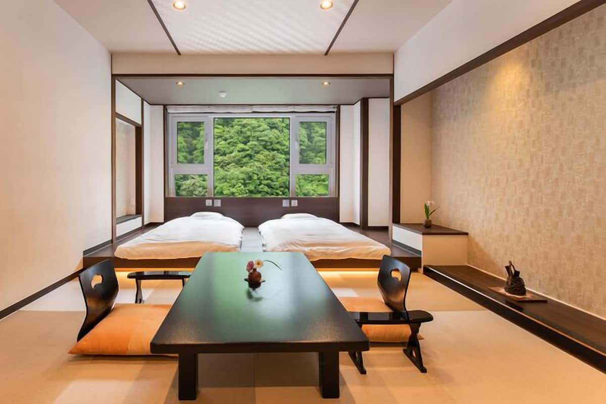 A room with two beds, a table, chairs, and a view of greenery through large windows.