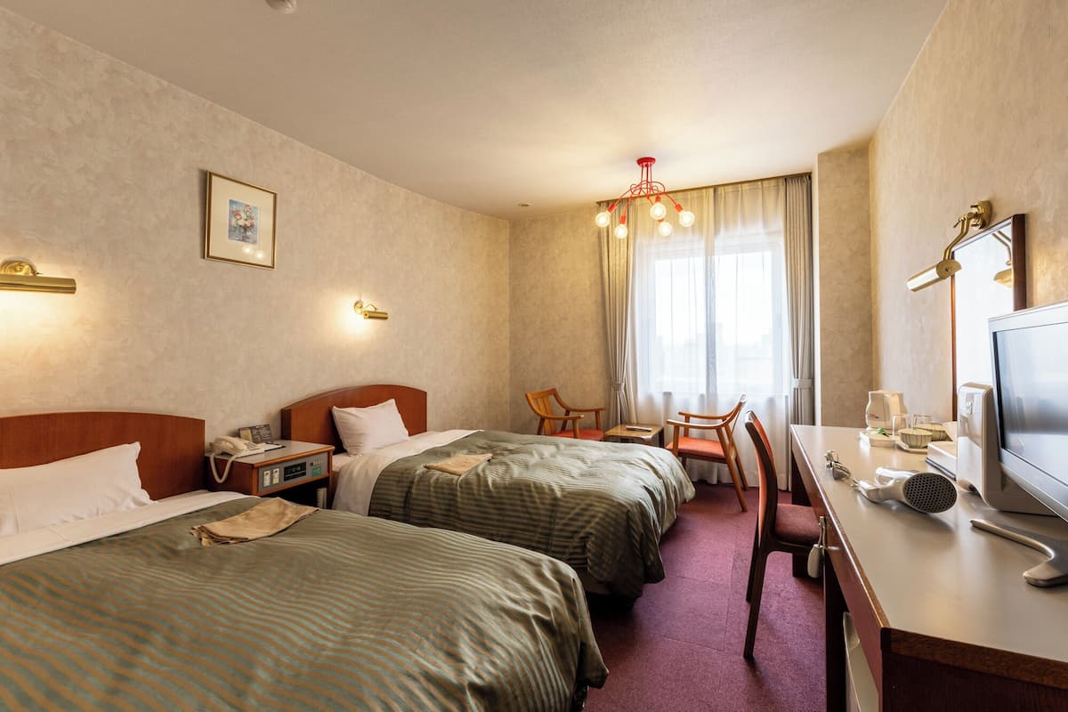 A hotel room with two beds, a desk with a TV, a chair, and a window with curtains.