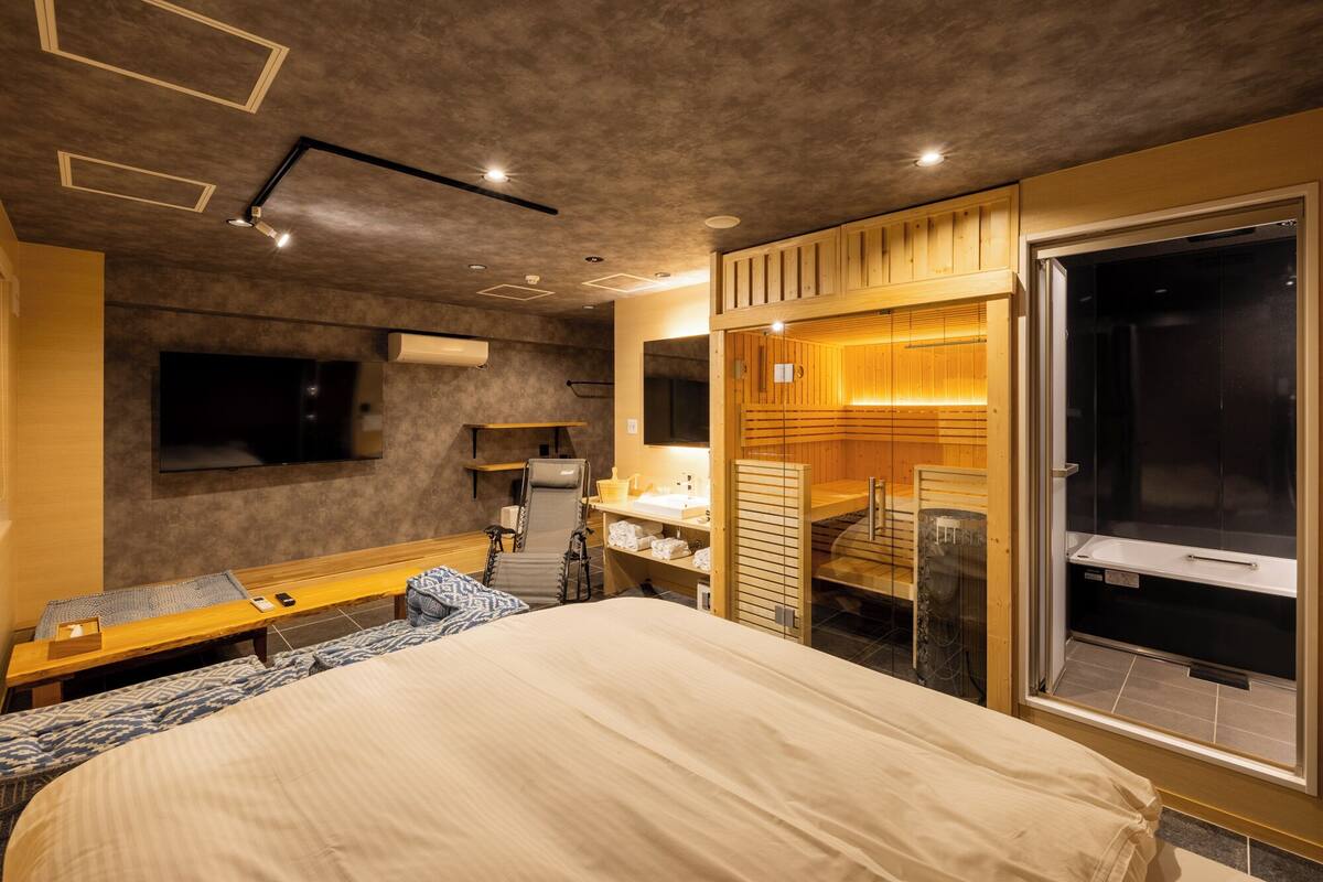 A modern hotel room with a large bed, a TV, a desk with a chair, and a sauna area.