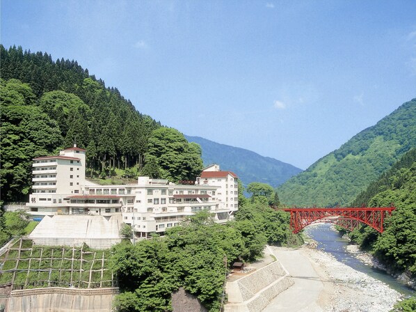 A multi-story hotel with a red bridge over a river, surrounded by lush greenery and mountains. - Hotel Kurobe (Kurobe)