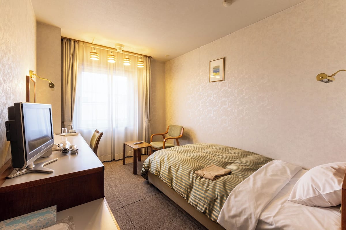 A hotel room with a bed, a desk with a computer, a chair, a window with curtains, and a wall-mounted light fixture.