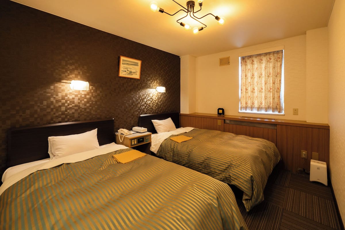 A hotel room with two beds, a nightstand with a phone, a clock, a window with curtains, and a wall-mounted light fixture.