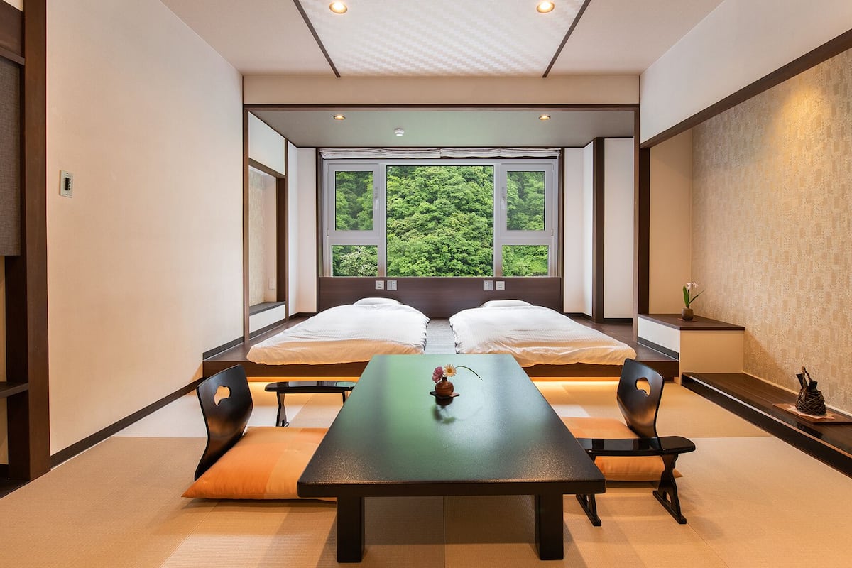 A room with two beds, a table, chairs, a window with a view of greenery, and a wall with a textured pattern.