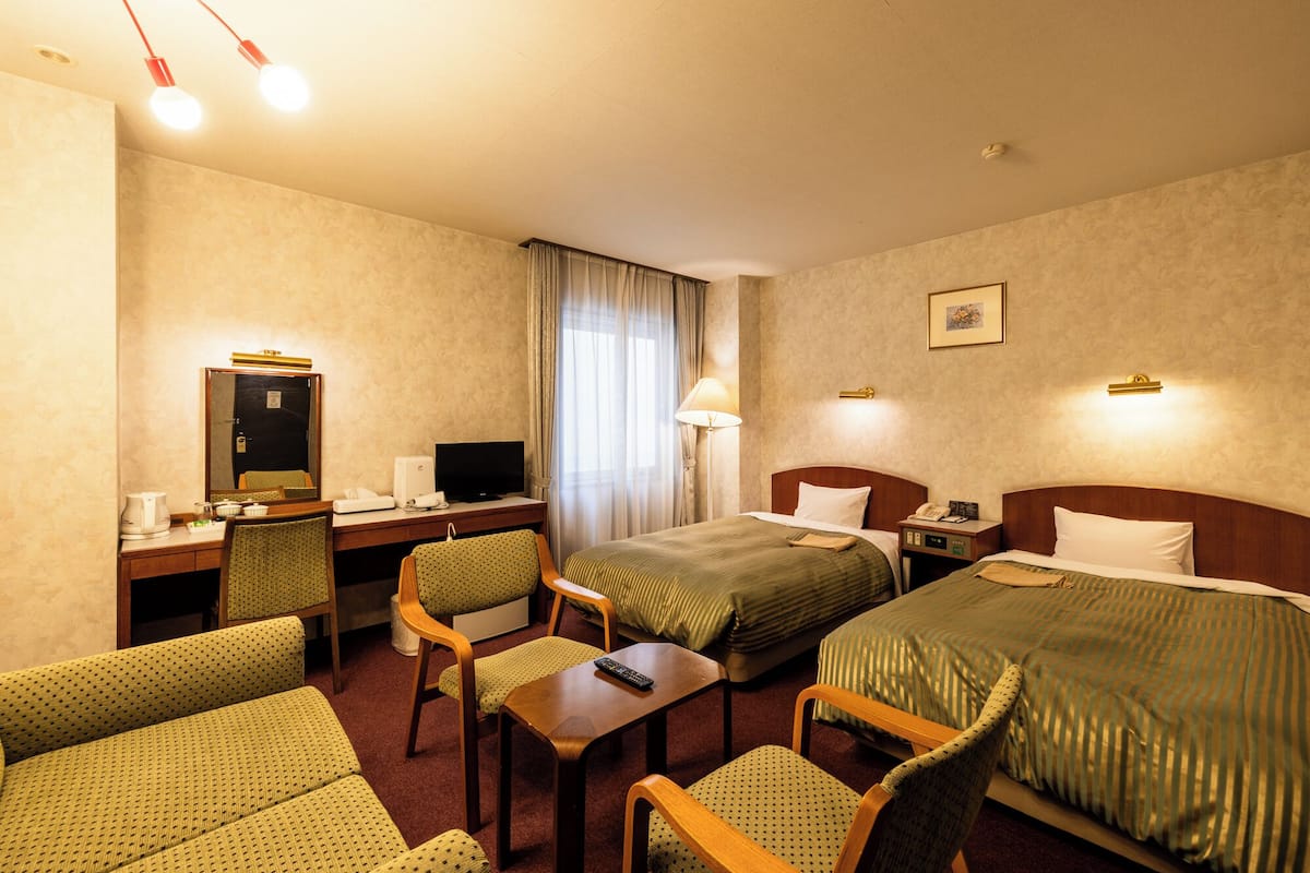 A hotel room with a desk, two beds, a chair, a TV, and a lamp.