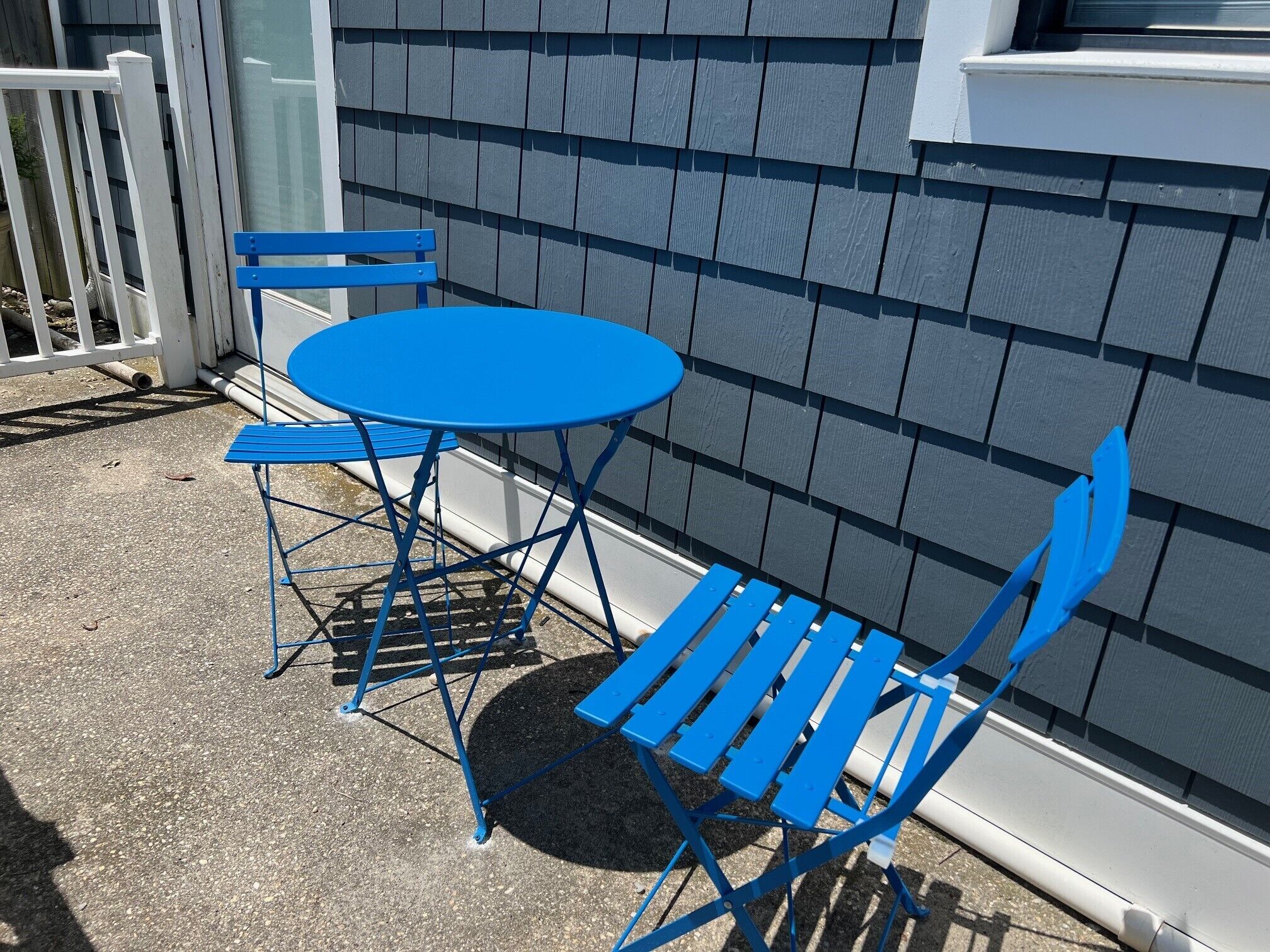 A blue metal table and chairs set on a concrete patio against a gray shingled wall.