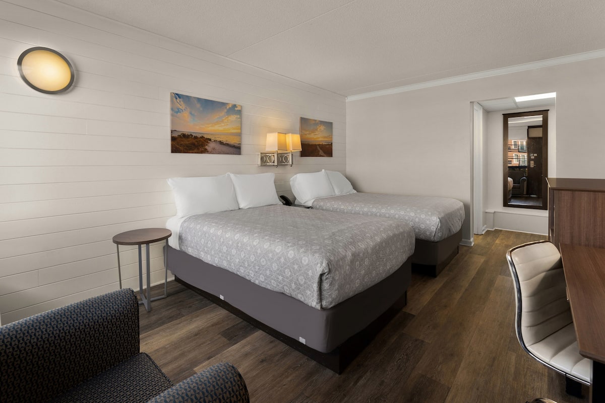 A hotel room with two beds, a small round table, a chair, and a desk. There are two wall-mounted lamps and two paintings above the beds.