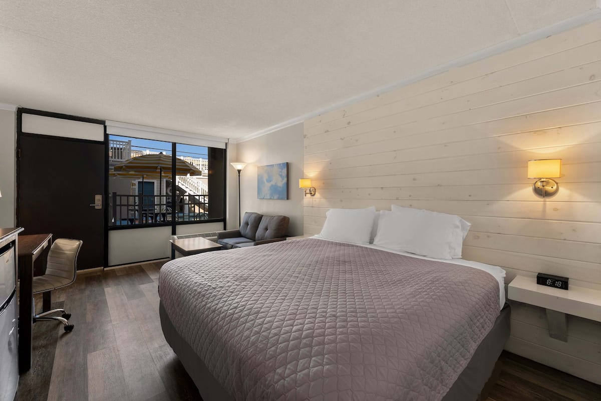 A hotel room with a large bed, a desk, and a chair. There is a window with a view of buildings outside.