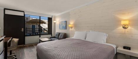 A hotel room with a large bed, a desk, and a chair. There is a window with a view of buildings outside.