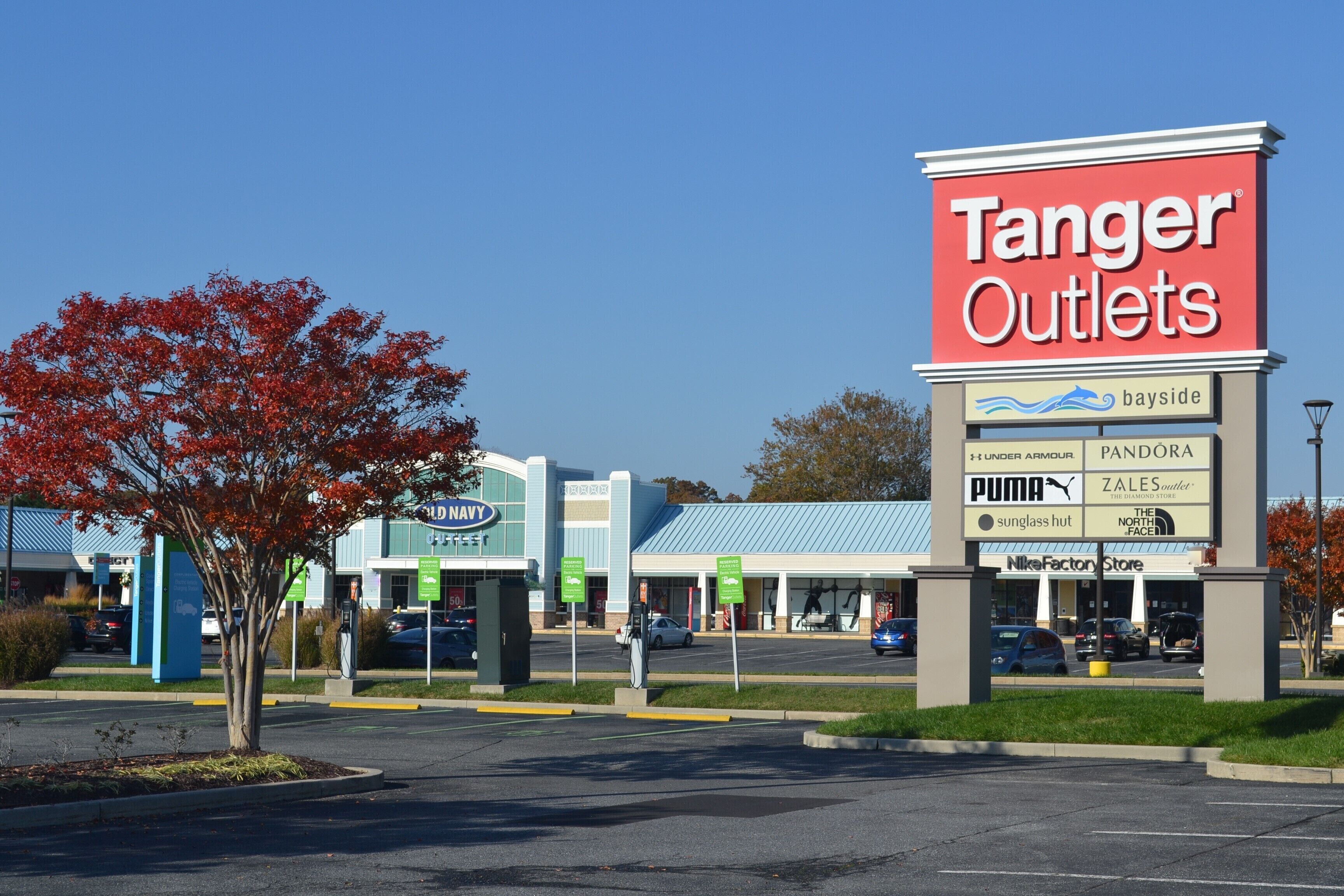Een bord van Tanger Outlets met diverse logo's van merken en een parkeerplaats.